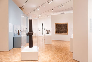 A view of a museum gallery shows a tall, narrow brown drum near the center. Beyond this and to the left is a glass case with carved figures. On a wall  slightly to the right of the drum, a large cloth in shades of brown is hangs in a frame on the wall.