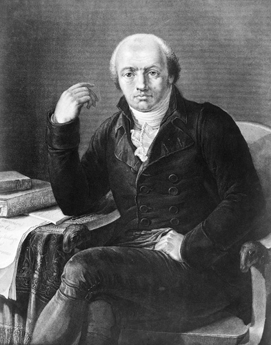 A black and white image of a seated man with crossed legs, a dark coat, a tall white collar, and a receding hairline. He leans on one elbow against a table covered in papers and books as he gazes directly out toward the viewer.