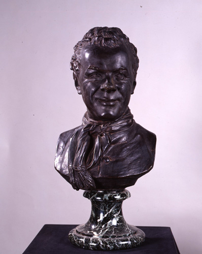A dark bronze portrait bust of a man with short, receding hair, lines at the corners of his eyes, and a faint smile. He has a scarf tied around his neck and looks away to the viewer's right.