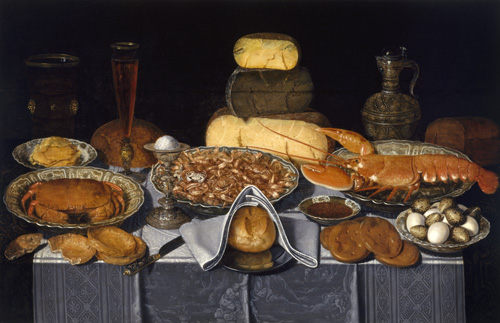 A table with a silver tablecloth is spread with food. The largest platters contain one crab, at the left, many shrimps, in the center, and one large lobster, on the right. In front of the seafood platters, three different kinds of baked breads or loaves are arranged, along with a bowl of eggs. Behind the seafood, a large stack of half-wheels of cheese is displayed, along with a glass wine vessel and a metal wine jug.