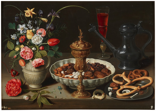 A table is set with food and flowers. On the left, a vase contains roses, daffodils, tulips, and other colorful flowers. Next to it, in the center of the image, a large white bowl contains dried fruit and nuts, with a gold-colored goblet in front of the bowl near the edge of the table. To the right of the bowl and goblet, a silver platter has several breadsticks and pretzels on it. Behind these, a glass containing red wine and a dark gray pitcher with a long spout are also shown.