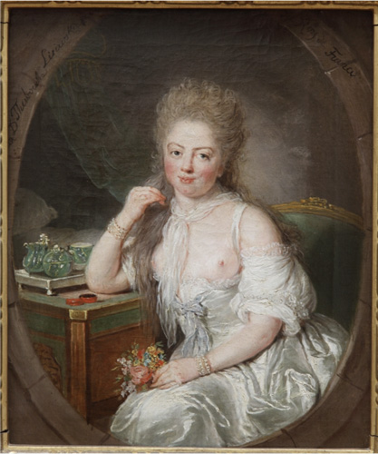 A woman with light skin, very pink cheeks, and long dark-blonde hair is seated at a small table with one elbow resting on it, gazing directly out at the viewer. She is wearing a silvery-white gown that is pulled below her breasts, along with a sheer white scarf tied around her neck and bracelets on both wrists. She holds a small bouquet of flowers in one hand.
