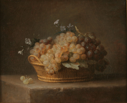 A shallow wicker basket with a handle is shown on a table. It has a large pile of grapes, some lighter in color and others dark purple, heaped inside, with several small flowers poking out .