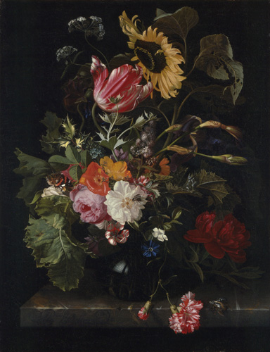 A dark vase of flowers sits on a table. The arrangement includes large green leaves, red and white carnations, a sunflower, roses, and other white, orange, yellow, and pink flowers against a dark background.