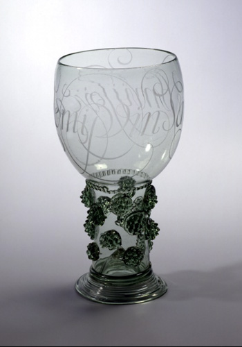 A glass goblet with raspberry-like glass shapes affixed all around the short stem of the glass. The bowl has an ornate, swirling script etched all around it.