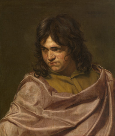 A young man with light skin, a round face, and shoulder-length, curly brown hair is shown from the chest up. He wears a mustard-yellow shirt beneath a reddish-brown cloak or wrap. He looks down and away from the viewer with a serious expression.