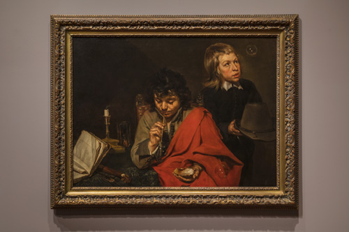 Two young boys with light skin, one with dark curly hair and one with straight blonde hair, are positioned near each other. The blonde child wears dark clothing and holds a hat, and is looking up at a soap bubble that is just above and to one side of his head. The dark-haired boy is seated, with a red cloth draped over one shoulder, holding a straw to his mouth and soap in the other hand. Nearby, a candle has gone out and a book fallen askew off the table.