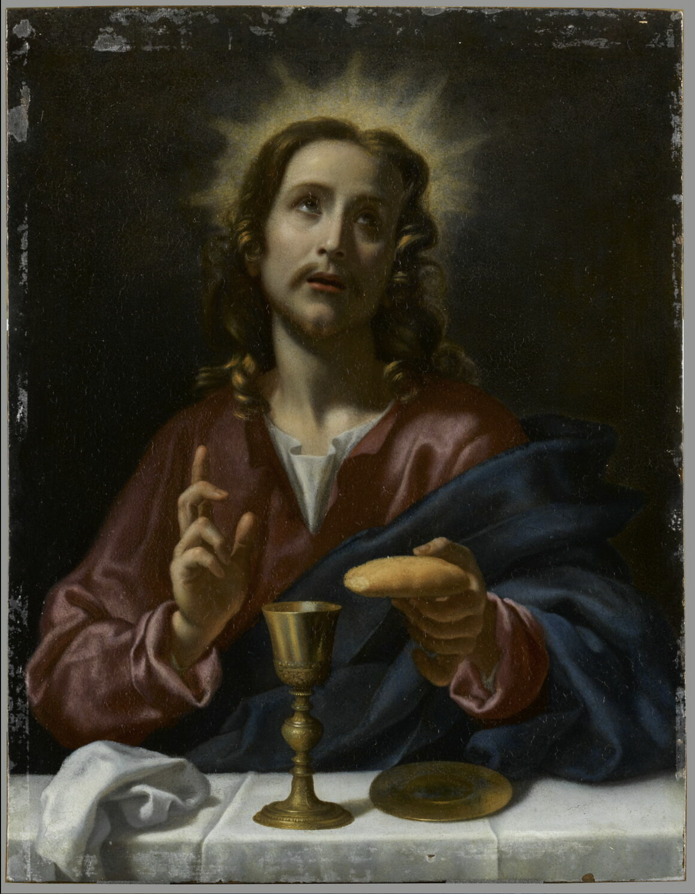 An image of Christ seated at a table with a gold goblet and a small gold plate in front of him. He is holding a piece of bread in his left hand, while his right hand is raised in blessing. He looks up towards the sky, while a halo of golden light frames his head.