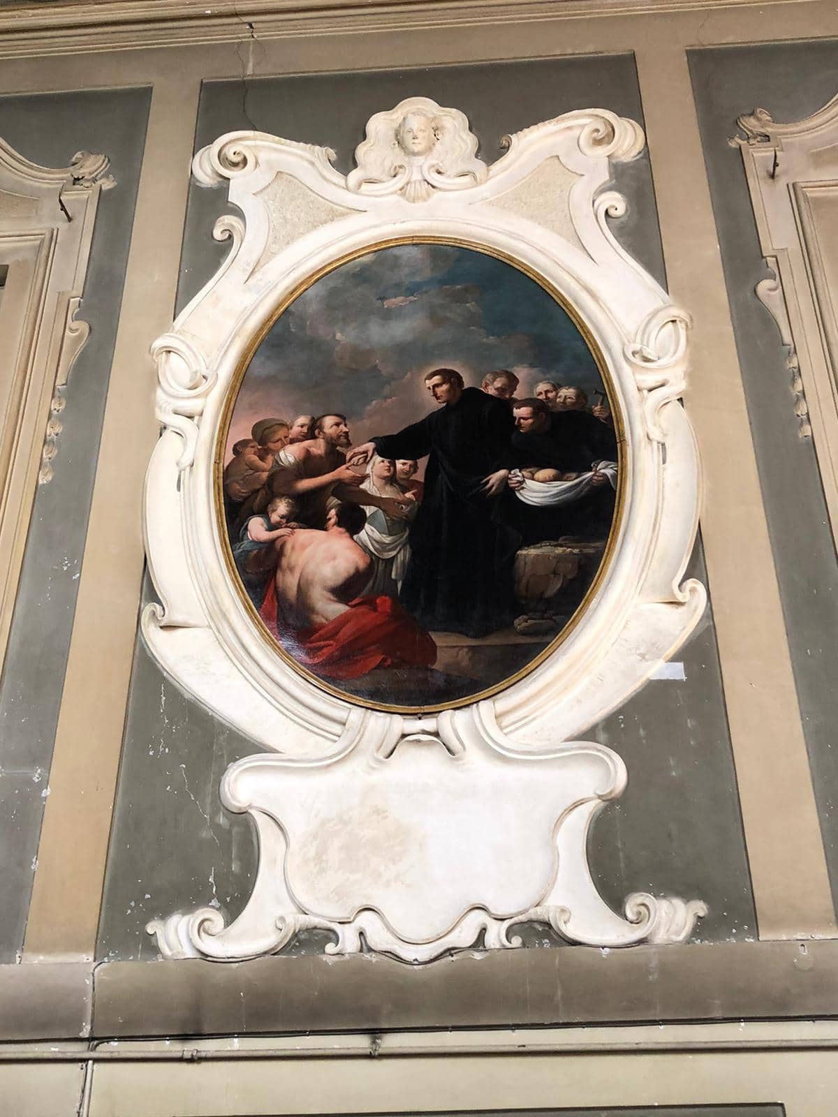 An oval painting in an elaborate marble frame, part of an architectural decoration. In the painting, a man in a long black robe hands bread to one of the many people gathered around him. Behind the group is a cloudy sky.