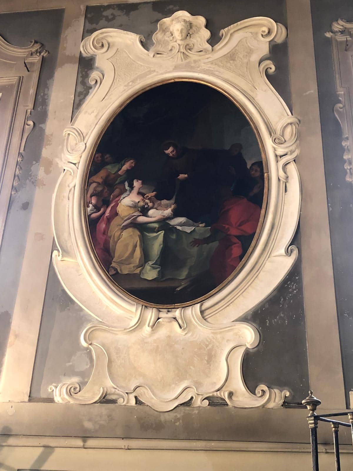 An oval painting in an elaborate marble frame, part of an architectural decoration. A man in a long black robe places his hand on the forehead of a sick child, who is being held by their mother. The mother and the other figures gathered around them look at the man with desperation.