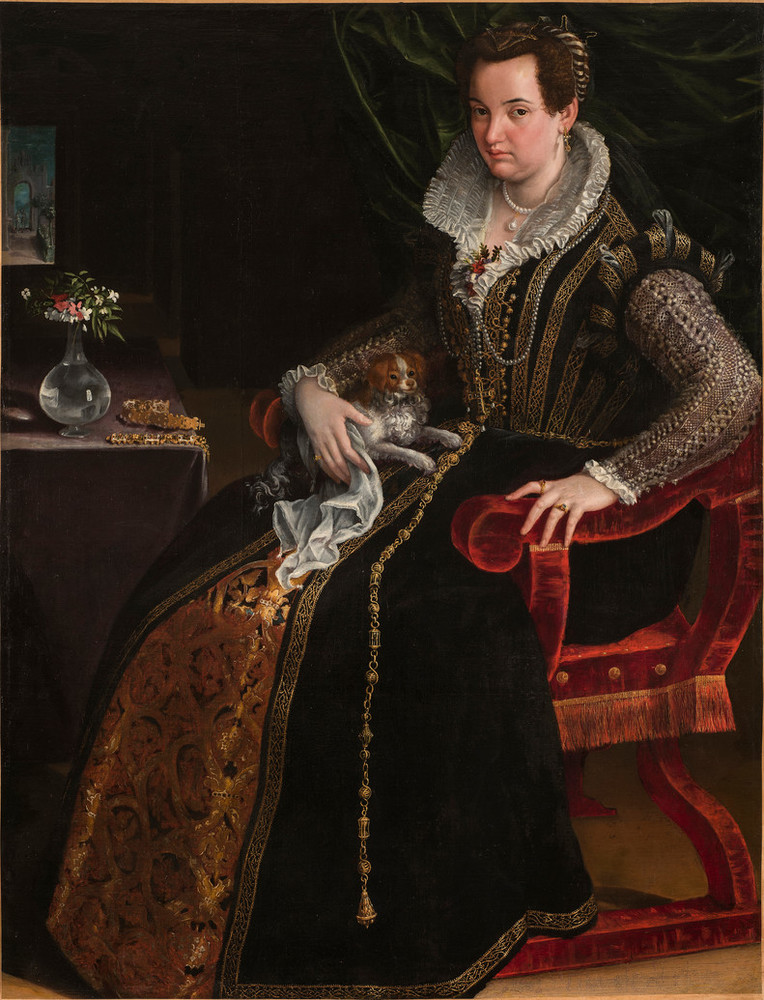 A full-length portrait of a woman seated in a red velvet chair. She is elegantly dressed in an elaborate sixteenth-century gown with a lace collar and embroidered details. A small white and brown dog sits on her lap, and she places her right hand on him. The table next to her has a small glass vase with flowers and some gold jewelry. 