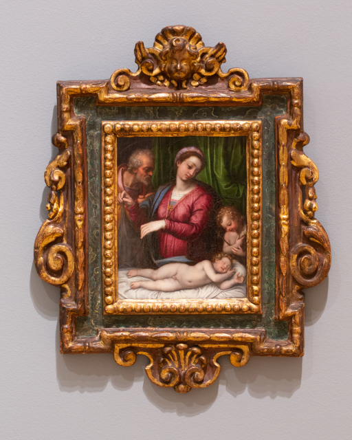 A small painting of the holy family in an ornate frame. The Infant Jesus is sleeping on his side while Mary and Joseph look down at him. The Infant Saint John the Baptist also looks down at him. The background is green drapery.