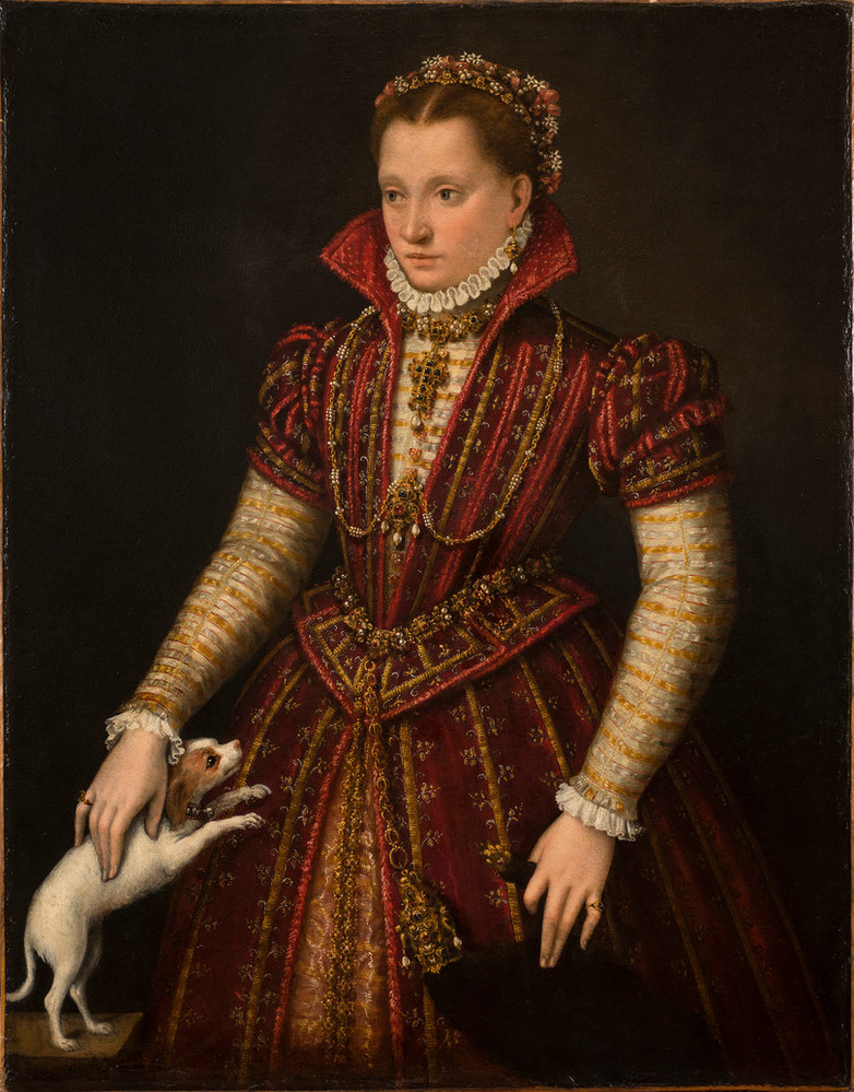 A half-length portrait of a woman in an ornate sixteenth-century gown, embellished with jewels. The gown is red, gold, and white, with a high collar and she wears a matching headband in her hair. Her right hand rests on a small brown and white dog that is standing on a small table to the side with its two front paws on the skirt of her gown.