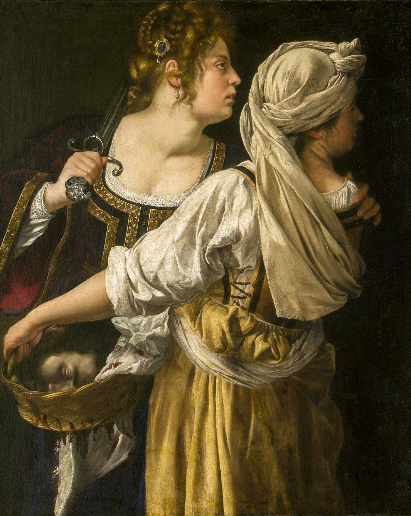Two women look off past the right edge of the canvas. They appear in half-length, and one woman faces us, holding a sword over her shoulder. She grips the shoulder of the other woman with her left hand. That woman wears a yellow dress and white head covering has her back to us and holds a basket to the side. Inside the basket is the bloodied head of a man. 
