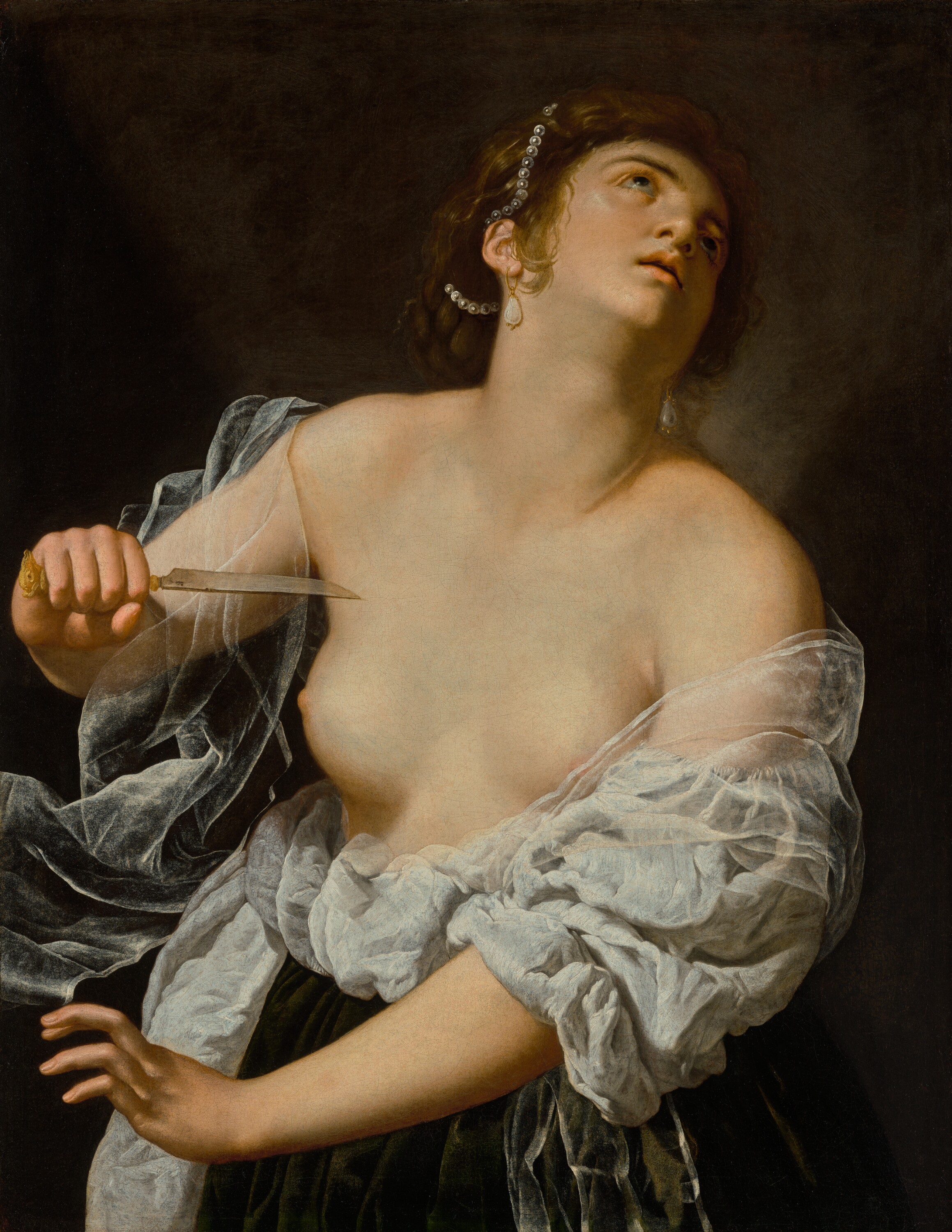 A half-length image of a woman in a diaphanous gown with both breasts bared. Her hair is pulled back from her face and adorned with pearls. She holds a knife in her right hand and aims the blade at her chest, looking up at the sky.