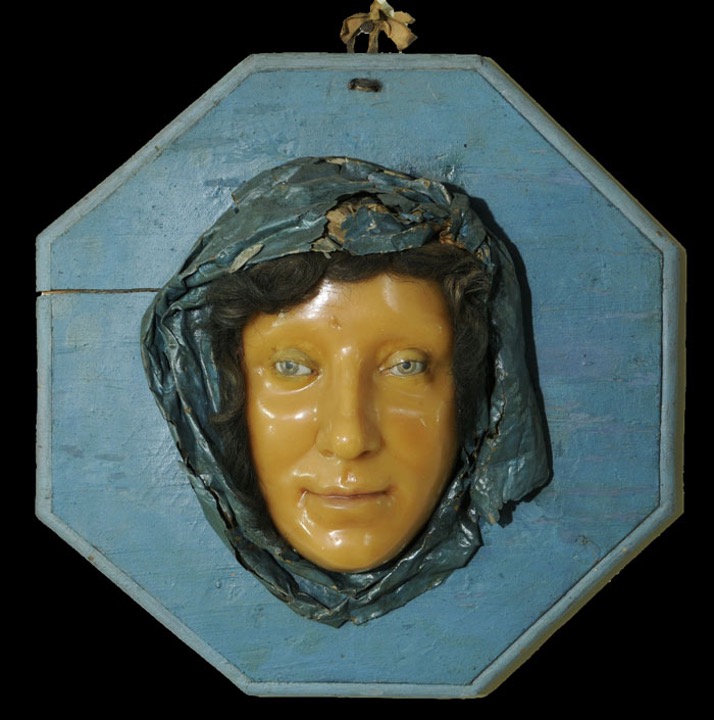 A portrait in relief of a woman’s face, surrounded by a blue head covering or veil. She looks out directly with a slight smile. 