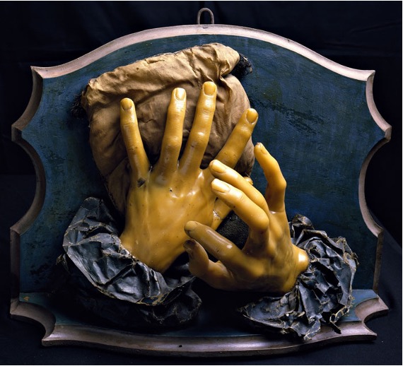 Two hands, modeled in relief and resting on top of one another. They are modeled in flesh-colored wax with blue sleeves visible below the wrists.
