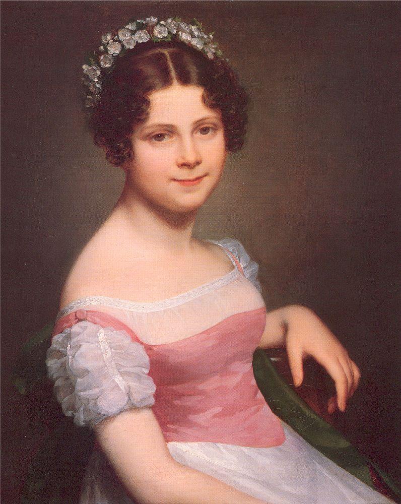 A half-length portrait of a young girl in a white and pink dress, sitting on a green chair. She has one elbow slung over the back of the chair, as she turns slightly. She wears a crown of flowers in her hair. She looks directly at the viewer with a closed mouth smile. 