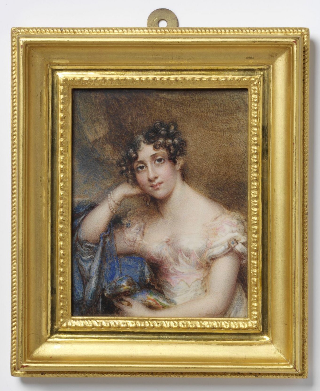 A half-length, rectangular portrait miniature of a young woman looking directly out at the viewer. She holds her head in one hand, with her elbow propped on a table covered in blue cloth. She wears two pearl bracelets and a pink gown, with her hair parted down the middle and her curls pulled up around her face.