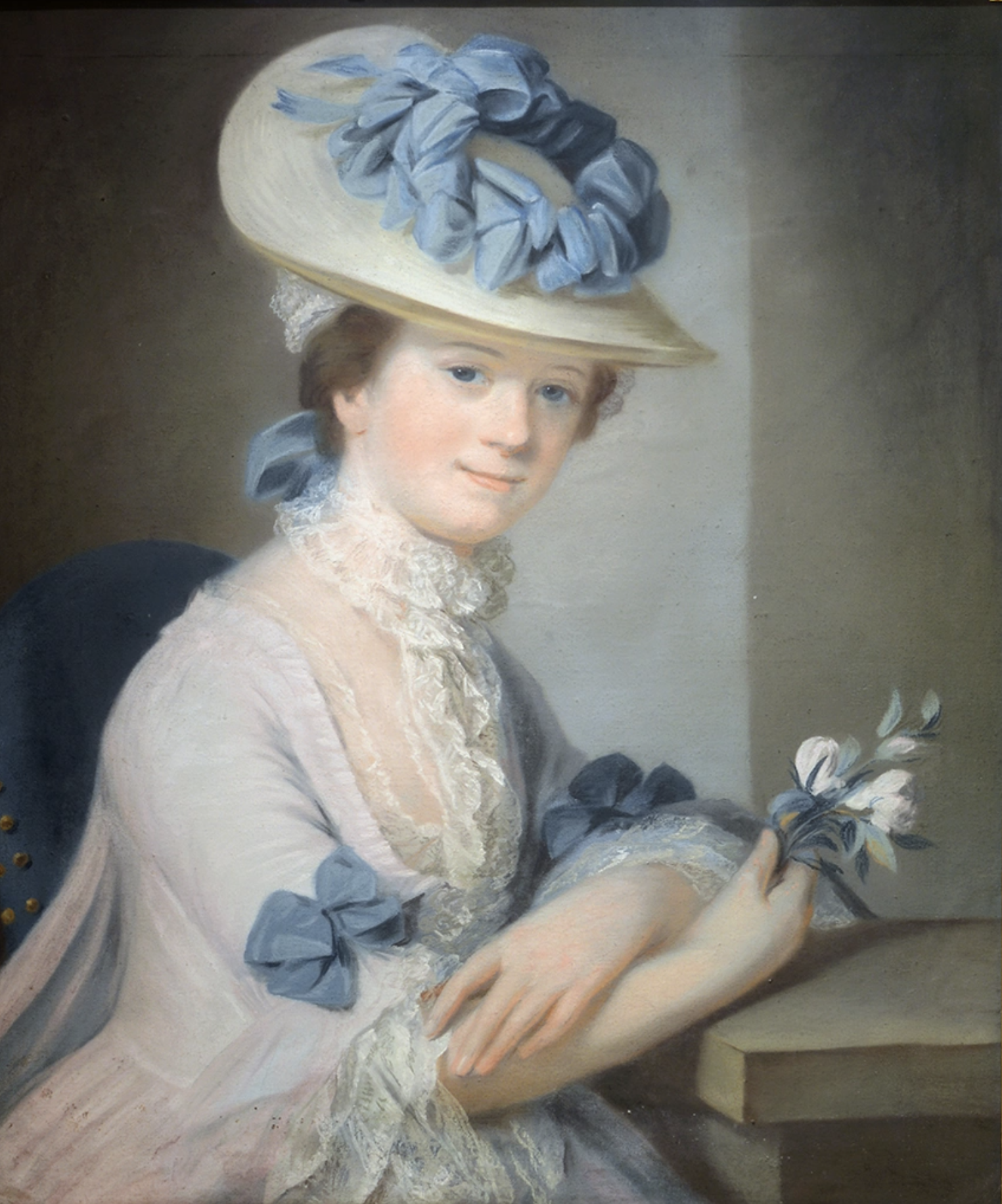 A half-length portrait of a young woman, seated on a blue chair and turned to meet the viewer’s gaze directly. She wears a cream dress with white lace and blue ribbons at her elbows, holding a sprig of white flowers in one hand. She wears a hat adorned with blue ribbons and bows, with more lace. Her expression is sweet, with a slight smile. 