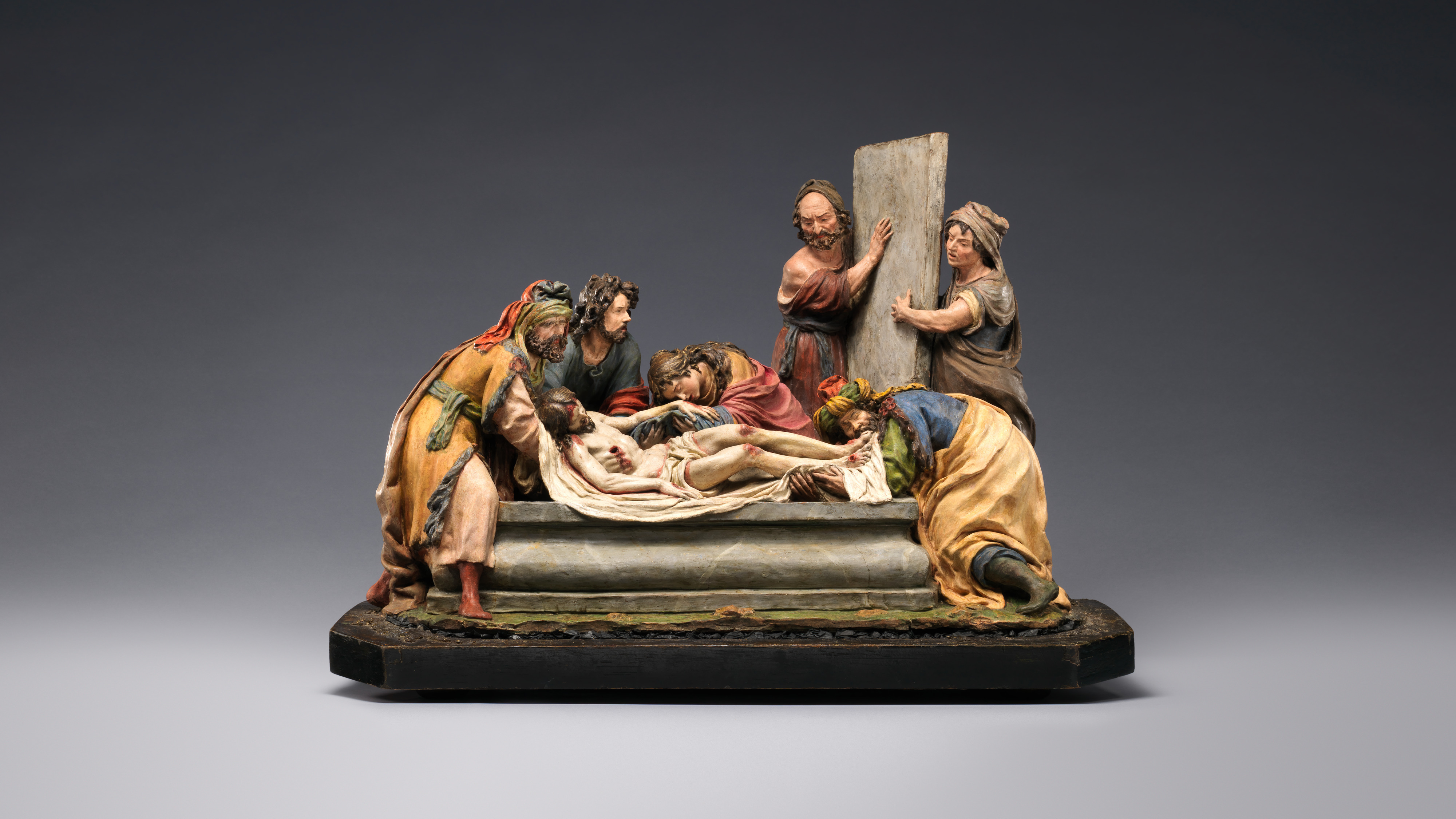 A polychrome terracotta sculpture of the entombment of Christ, who is being laid in a stone tomb by several figures. Two men hold the tomb’s cover while a third figure kisses his feet. A woman rests her face on his hand, in anguish. Two other men lift him by the shoulders. Christ is nude except for a draped cloth, which leaves the wounds of his crucifixion visible.