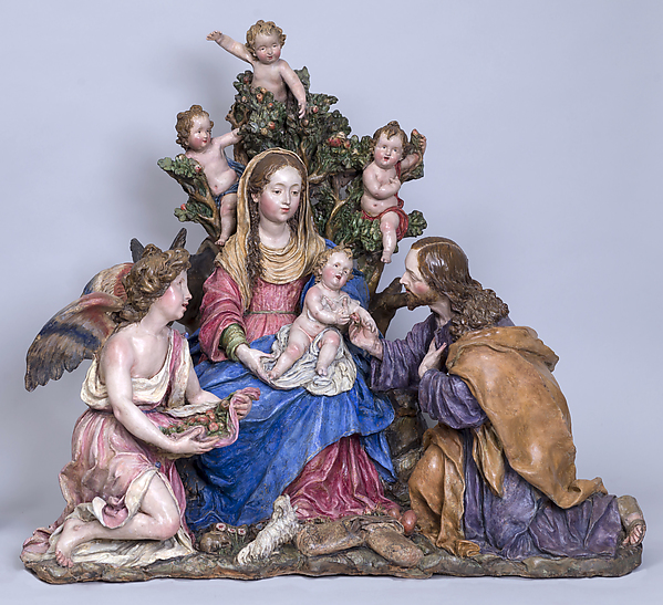 A polychrome terracotta sculpture of the Virgin Mary seated in a tree, holding the infant Jesus in her lap. Joseph kneels beside her, holding Jesus’s hand. An angel kneels to her right holding flowers, while three putti figures are seated in the tree behind Mary. Her expression is neutral and serene, and she looks away from the viewer. 