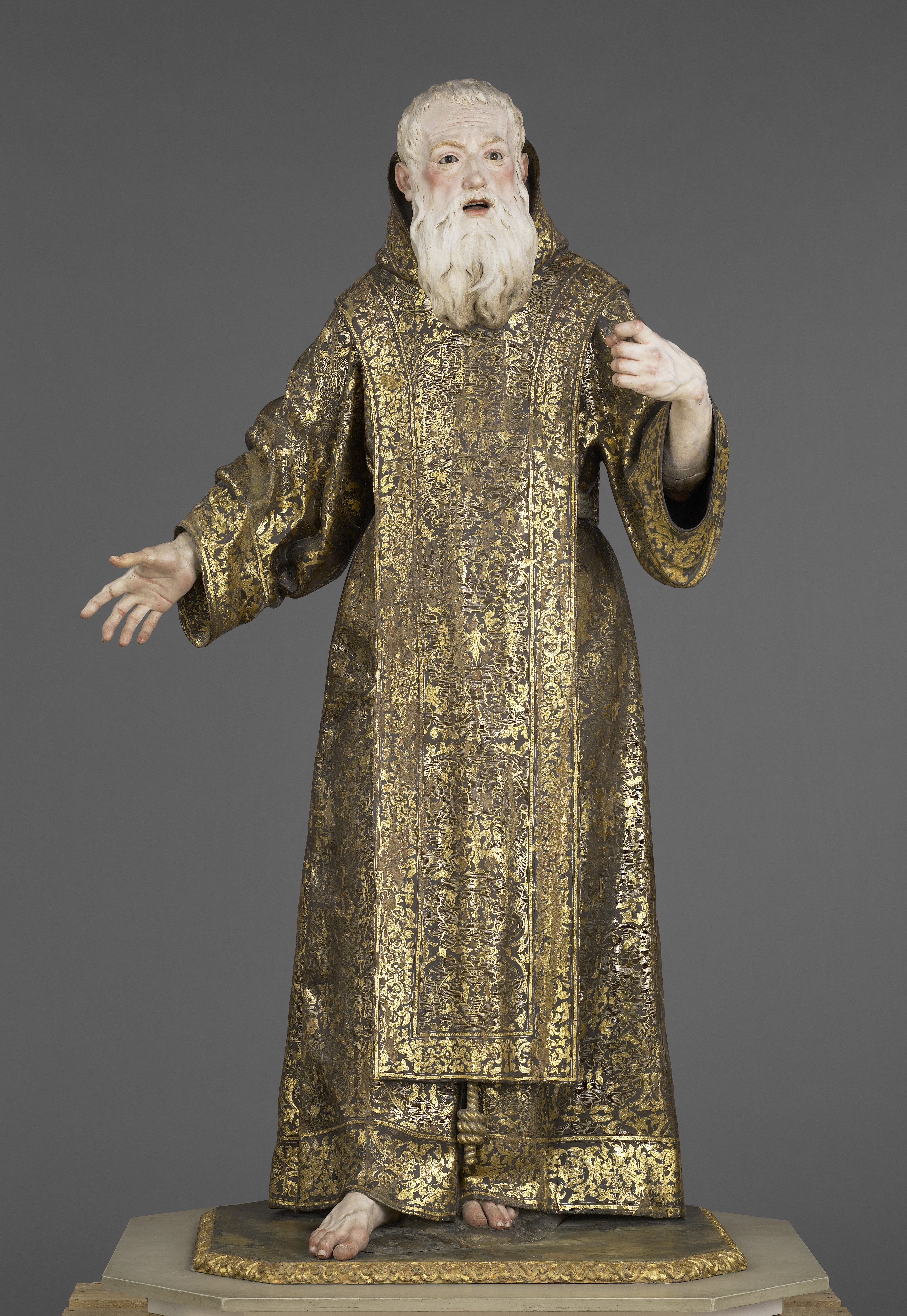 A polychrome wood sculpture of an older man standing, wearing a long gold embellished robe with a hood. He has white hair and a long white beard and he holds out his hands as though he is speaking. His lips are parted and his eyebrows are raised. Despite his elaborate robe, he is barefoot.