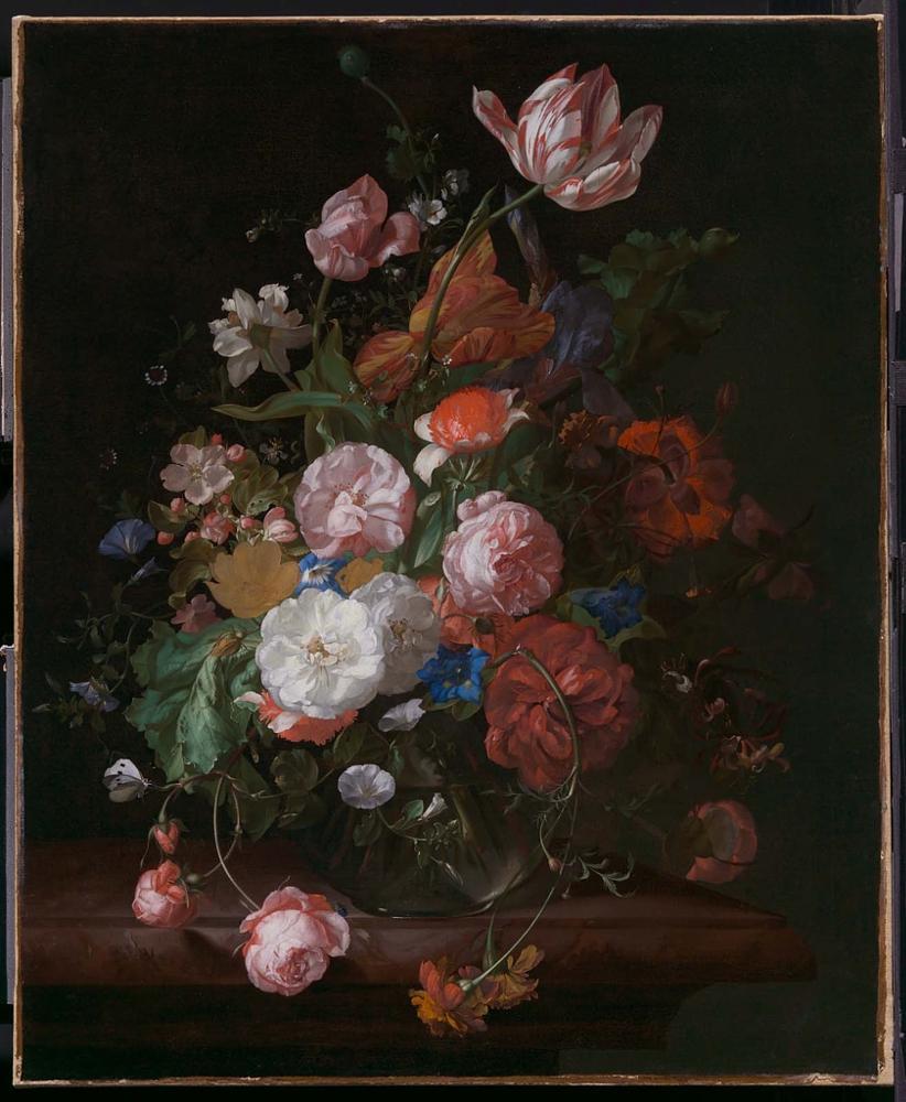 A floral still life set against a dark background. The arrangement is sitting on a brown ledge. The flowers are colorful: pink, red, blue, white, and orange, as well as lush green leaves. 
