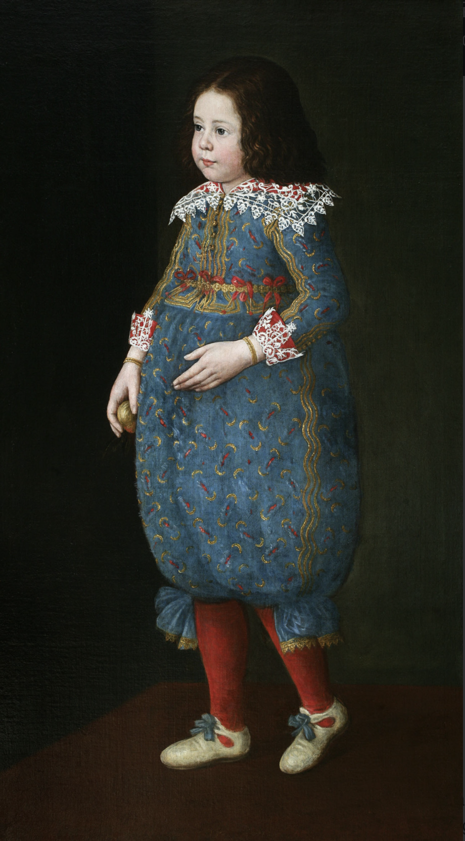 A full-length portrait of a boy wearing an elaborate costume that includes embroidered blue pantaloons with red stockings. He has lace on his collar and at his sleeves. He has long, curly hair past his shoulders. The background is dark and neutral, and the boy looks off to the left side of the canvas.