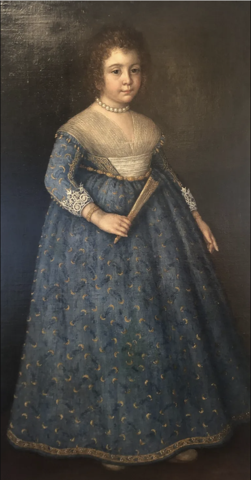 A full-length portrait of a girl wearing a blue dress with a sheer detail at the bust and lace at her sleeves. She wears a pearl necklace and has her curly brown hair pulled back. She holds a closed fan in her right hand. The background behind her is dark and neutral.